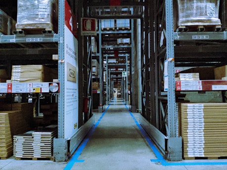 A warehouse aisle lined with tall metal shelves stacked with packaged goods. The shelves have labels and signs indicating different sections and product codes. The floor guides with blue lines direct the traffic flow within the facility.