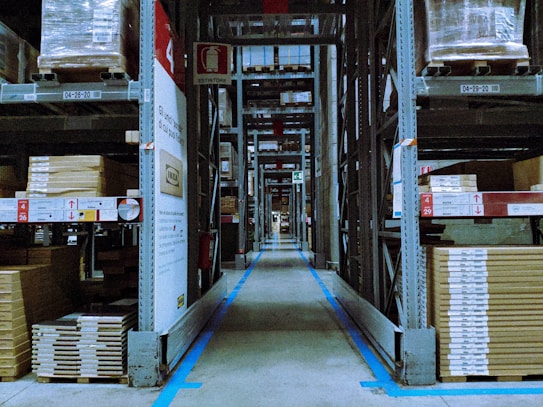 A warehouse aisle lined with tall metal shelves stacked with packaged goods. The shelves have labels and signs indicating different sections and product codes. The floor guides with blue lines direct the traffic flow within the facility.