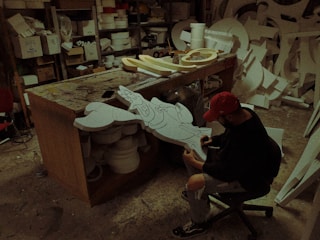 A person wearing a red cap and black clothing is sitting on a chair and working on a cartoon character cut-out in a workshop. The workshop is cluttered with various materials, tools, and other cut-outs. The person appears to be drawing or painting the cut-out piece.
