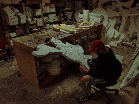 A person wearing a red cap and black clothing is sitting on a chair and working on a cartoon character cut-out in a workshop. The workshop is cluttered with various materials, tools, and other cut-outs. The person appears to be drawing or painting the cut-out piece.