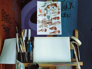 An artistic workspace featuring a wooden easel with a paper displaying sketches of lips, surrounded by various brushes and art tools. The walls have graffiti and abstract designs, adding an urban and creative atmosphere.