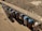 A row of spray paint cans is lined up along a concrete curb. The ground is covered in gravel and the lighting casts shadows from the cans onto the pavement. Each can has a blue cap, and various colors and designs are displayed on the labels.
