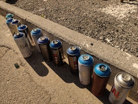 A row of spray paint cans is lined up along a concrete curb. The ground is covered in gravel and the lighting casts shadows from the cans onto the pavement. Each can has a blue cap, and various colors and designs are displayed on the labels.