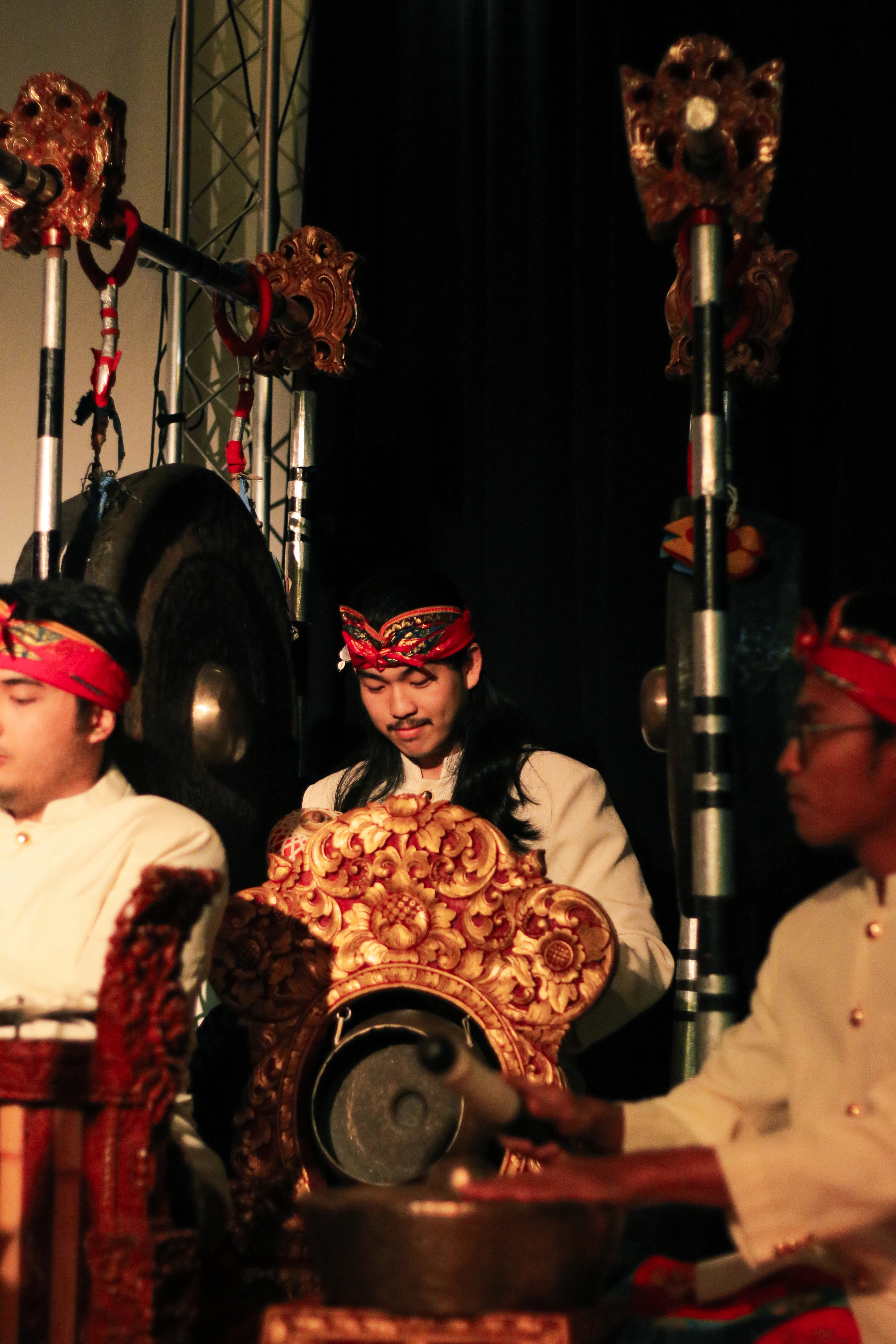 Gamelan musicians in red headscarves play gold-carved percussion instruments under dramatic stage lighting.