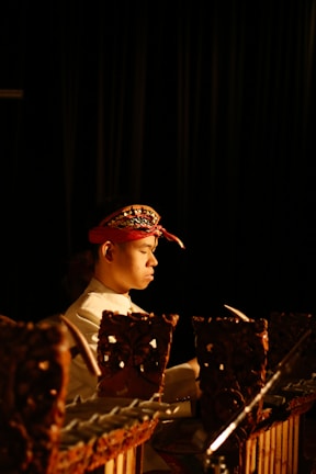 A person is playing a traditional wooden musical instrument, wearing a red headband with intricate designs. The setting is dimly lit, creating a focused and intimate atmosphere.