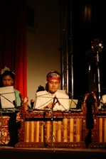 A man in traditional attire plays a wooden xylophone-like musical instrument. He is seated in front of a music stand holding sheets. The scene is dimly lit, creating a focused spotlight on the musician, enhancing the cultural and performance ambiance.