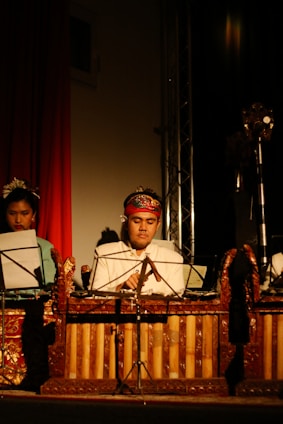 A man in traditional attire plays a wooden xylophone-like musical instrument. He is seated in front of a music stand holding sheets. The scene is dimly lit, creating a focused spotlight on the musician, enhancing the cultural and performance ambiance.