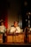 A man in traditional attire plays a wooden xylophone-like musical instrument. He is seated in front of a music stand holding sheets. The scene is dimly lit, creating a focused spotlight on the musician, enhancing the cultural and performance ambiance.