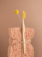 A natural sponge with a rough texture is tied around the middle with twine. An eco-friendly bamboo toothbrush with yellow bristles and a single yellow billy button flower are nestled into the sponge. The background is a soft beige color, complementing the neutral tones of the sponge and toothbrush.