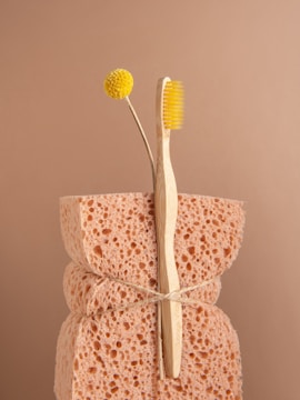 A natural sponge with a rough texture is tied around the middle with twine. An eco-friendly bamboo toothbrush with yellow bristles and a single yellow billy button flower are nestled into the sponge. The background is a soft beige color, complementing the neutral tones of the sponge and toothbrush.
