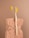 A natural sponge with a rough texture is tied around the middle with twine. An eco-friendly bamboo toothbrush with yellow bristles and a single yellow billy button flower are nestled into the sponge. The background is a soft beige color, complementing the neutral tones of the sponge and toothbrush.