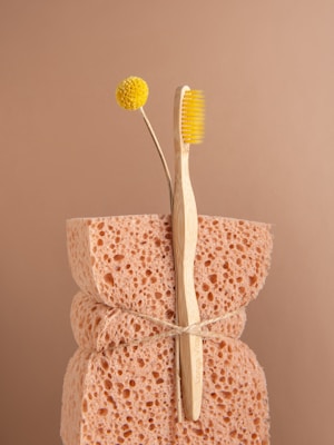 A natural sponge with a rough texture is tied around the middle with twine. An eco-friendly bamboo toothbrush with yellow bristles and a single yellow billy button flower are nestled into the sponge. The background is a soft beige color, complementing the neutral tones of the sponge and toothbrush.