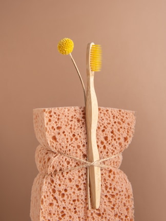 A natural sponge with a rough texture is tied around the middle with twine. An eco-friendly bamboo toothbrush with yellow bristles and a single yellow billy button flower are nestled into the sponge. The background is a soft beige color, complementing the neutral tones of the sponge and toothbrush.