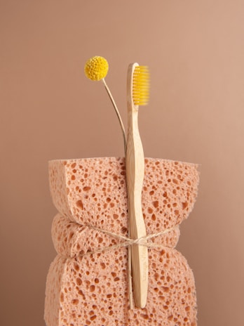 A natural sponge with a rough texture is tied around the middle with twine. An eco-friendly bamboo toothbrush with yellow bristles and a single yellow billy button flower are nestled into the sponge. The background is a soft beige color, complementing the neutral tones of the sponge and toothbrush.