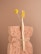 A natural sponge with a rough texture is tied around the middle with twine. An eco-friendly bamboo toothbrush with yellow bristles and a single yellow billy button flower are nestled into the sponge. The background is a soft beige color, complementing the neutral tones of the sponge and toothbrush.