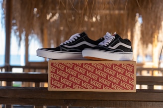 A pair of black and white sneakers rests on a branded shoebox placed on a wooden railing. The background features a thatched roof with sunlight filtering through, and a glimpse of the ocean in the distance.