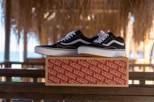 A pair of black and white sneakers rests on a branded shoebox placed on a wooden railing. The background features a thatched roof with sunlight filtering through, and a glimpse of the ocean in the distance.
