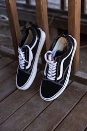 A pair of black and white sneakers with white laces is leaning against a wooden railing on a deck. The shoes have a minimalist design with the brand's logo visible inside.
