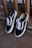A pair of black and white sneakers with white laces is leaning against a wooden railing on a deck. The shoes have a minimalist design with the brand's logo visible inside.