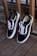 A pair of black and white sneakers with white laces is leaning against a wooden railing on a deck. The shoes have a minimalist design with the brand's logo visible inside.