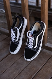 A pair of black and white sneakers with white laces is leaning against a wooden railing on a deck. The shoes have a minimalist design with the brand's logo visible inside.