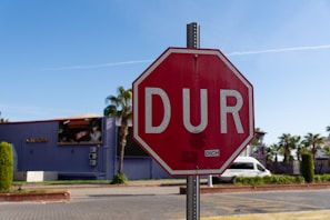 A red octagonal stop sign with the word 'DUR' in white letters is prominently displayed in the foreground. The background features a street scene with a blue building, palm trees, and a parked white van under a clear blue sky.