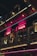 A dimly lit restaurant exterior with pink neon signs displaying words related to Peruvian cuisine. The scene includes a balcony with glass railings where people are walking. The building has an industrial design with metal and wood elements. Hanging flower baskets are visible, adding a touch of greenery.