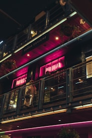 A dimly lit restaurant exterior with pink neon signs displaying words related to Peruvian cuisine. The scene includes a balcony with glass railings where people are walking. The building has an industrial design with metal and wood elements. Hanging flower baskets are visible, adding a touch of greenery.