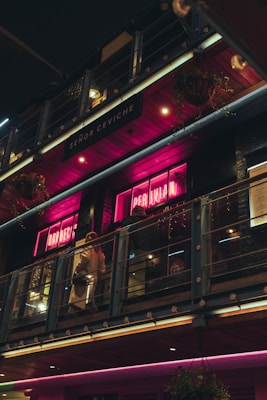 A dimly lit restaurant exterior with pink neon signs displaying words related to Peruvian cuisine. The scene includes a balcony with glass railings where people are walking. The building has an industrial design with metal and wood elements. Hanging flower baskets are visible, adding a touch of greenery.