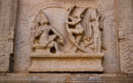 An intricately carved stone bas-relief depicting a mythological scene with two human figures. One figure is seated with legs crossed, possibly a deity, adorned in traditional attire and a headdress. The other figure appears dynamic, possibly engaged in a dance or combat pose, holding a bow. The carving is surrounded by ornate engravings on a weathered stone surface, suggesting historical or religious significance.