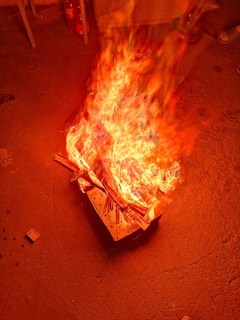 A vivid fire is burning intensely on a surface surrounded by various objects. The flames are prominently orange and yellow, indicating a strong heat and energy. Nearby, there are some containers and a matchbox on the ground, indicating the fire may have been deliberately started.