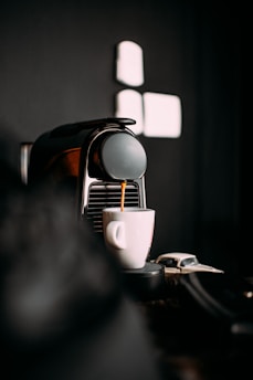 A sleek espresso machine dispensing coffee into a white cup. The background is dark, creating a stark contrast that highlights the coffee-making process.