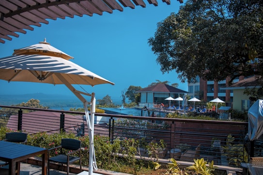 A serene outdoor setting featuring a patio with chairs and tables under large parasols. In the background, a swimming pool area is visible with more parasols and lounge chairs. The scene is complemented by lush greenery and distant hills beneath a clear blue sky.