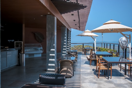 An outdoor dining area with several tables and chairs under large umbrellas. The setting appears to be on a balcony or terrace with a view of the sea and distant hills under a clear blue sky. The design incorporates modern elements with glass and metal railings.