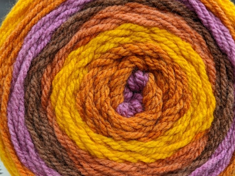 A close-up view of a rolled-up skein of yarn featuring concentric rings of different colors. The yarn is tightly wound, showcasing a vibrant pattern with distinct layers of color.