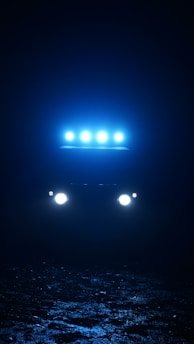 A vehicle with bright headlights and a series of roof lights glowing in a dark environment. The ground appears rough and textured, reflecting some of the light.