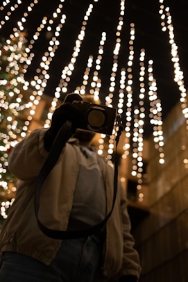 A person is seen holding a camera under a canopy of warm, glowing string lights against a dark background, creating a festive and cozy atmosphere.