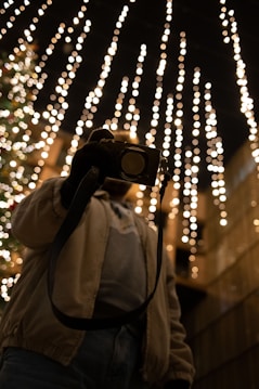 A person is seen holding a camera under a canopy of warm, glowing string lights against a dark background, creating a festive and cozy atmosphere.
