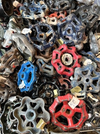 A collection of assorted vintage faucet handles in various shapes and colors, including blue, red, black, and gray, piled together. Each handle has a distinct design and some have visible price tags attached.