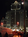 A tall, illuminated hotel building at night with a sign that reads 'RIU' at the top. The facade is well-lit, showcasing numerous windows. In the foreground, there are constructions and a crowd of people walking. Bright streetlights and festive lights add a warm glow to the scene.