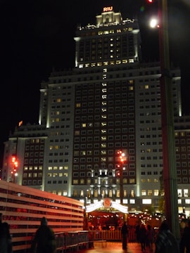 A tall, illuminated hotel building at night with a sign that reads 'RIU' at the top. The facade is well-lit, showcasing numerous windows. In the foreground, there are constructions and a crowd of people walking. Bright streetlights and festive lights add a warm glow to the scene.