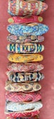 A collection of vibrantly colored, intricately patterned shoes arranged in a vertical line on a textured red surface. Each shoe features unique designs with floral, geometric, and abstract patterns in various bright colors.