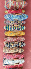 A collection of vibrantly colored, intricately patterned shoes arranged in a vertical line on a textured red surface. Each shoe features unique designs with floral, geometric, and abstract patterns in various bright colors.