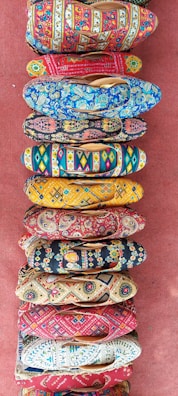 A collection of vibrantly colored, intricately patterned shoes arranged in a vertical line on a textured red surface. Each shoe features unique designs with floral, geometric, and abstract patterns in various bright colors.