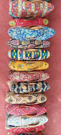 A collection of vibrantly colored, intricately patterned shoes arranged in a vertical line on a textured red surface. Each shoe features unique designs with floral, geometric, and abstract patterns in various bright colors.