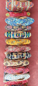 A collection of vibrantly colored, intricately patterned shoes arranged in a vertical line on a textured red surface. Each shoe features unique designs with floral, geometric, and abstract patterns in various bright colors.