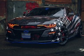 A sleek sports car with a bold American flag theme covering its body. The vehicle features distinctive red headlights, a glossy finish, and prominent star and striped patterns. It's parked in an urban environment, adding to its edgy appearance.