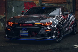 A sleek sports car with a bold American flag theme covering its body. The vehicle features distinctive red headlights, a glossy finish, and prominent star and striped patterns. It's parked in an urban environment, adding to its edgy appearance.