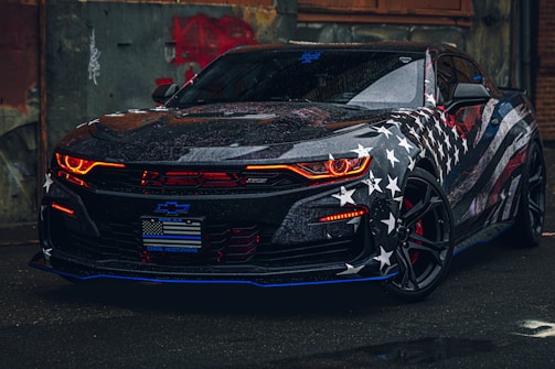 A sleek sports car with a bold American flag theme covering its body. The vehicle features distinctive red headlights, a glossy finish, and prominent star and striped patterns. It's parked in an urban environment, adding to its edgy appearance.