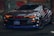 A sleek sports car with a bold American flag theme covering its body. The vehicle features distinctive red headlights, a glossy finish, and prominent star and striped patterns. It's parked in an urban environment, adding to its edgy appearance.